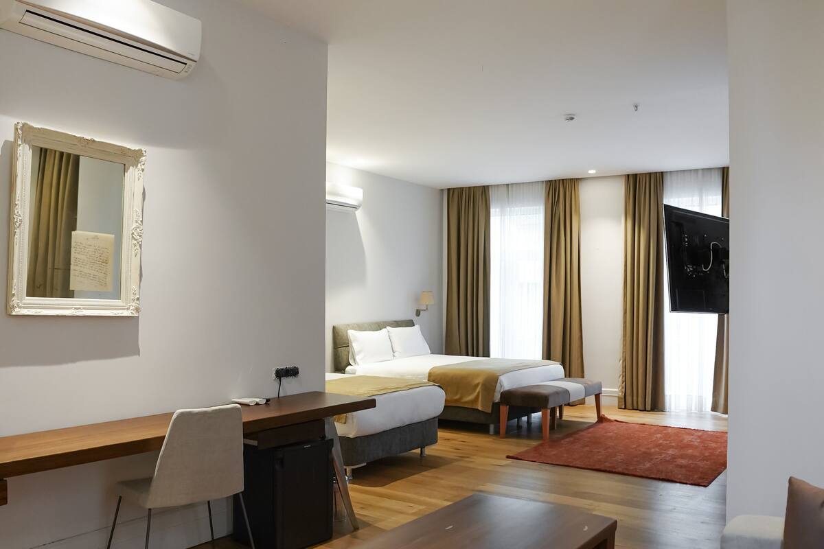 A modern hotel room with a wooden desk, a bed with a beige bedspread, a sofa, and a TV.