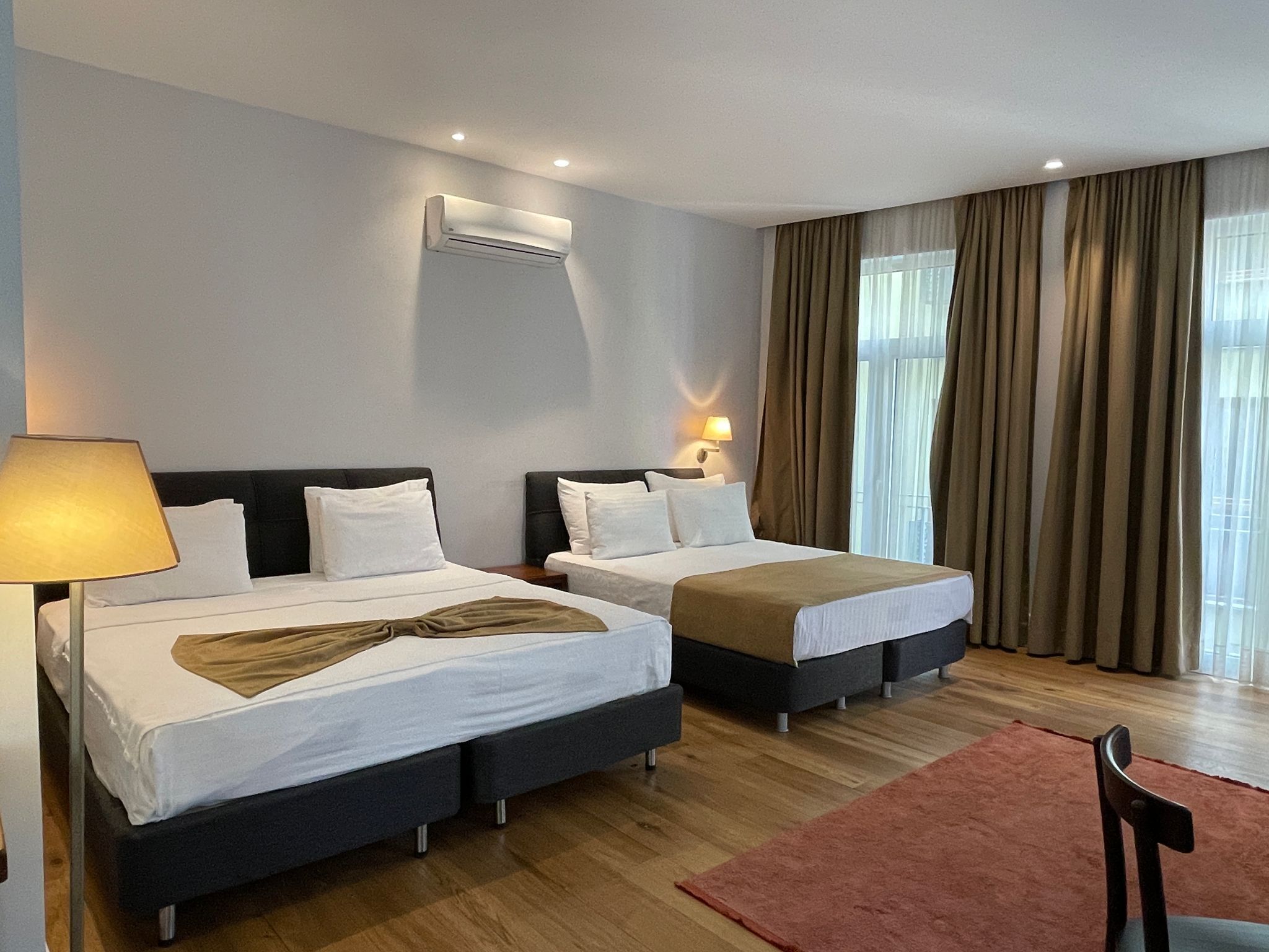 A hotel room with two beds, a wall-mounted air conditioner, a bedside lamp, and a view of the outdoors through large windows with curtains.