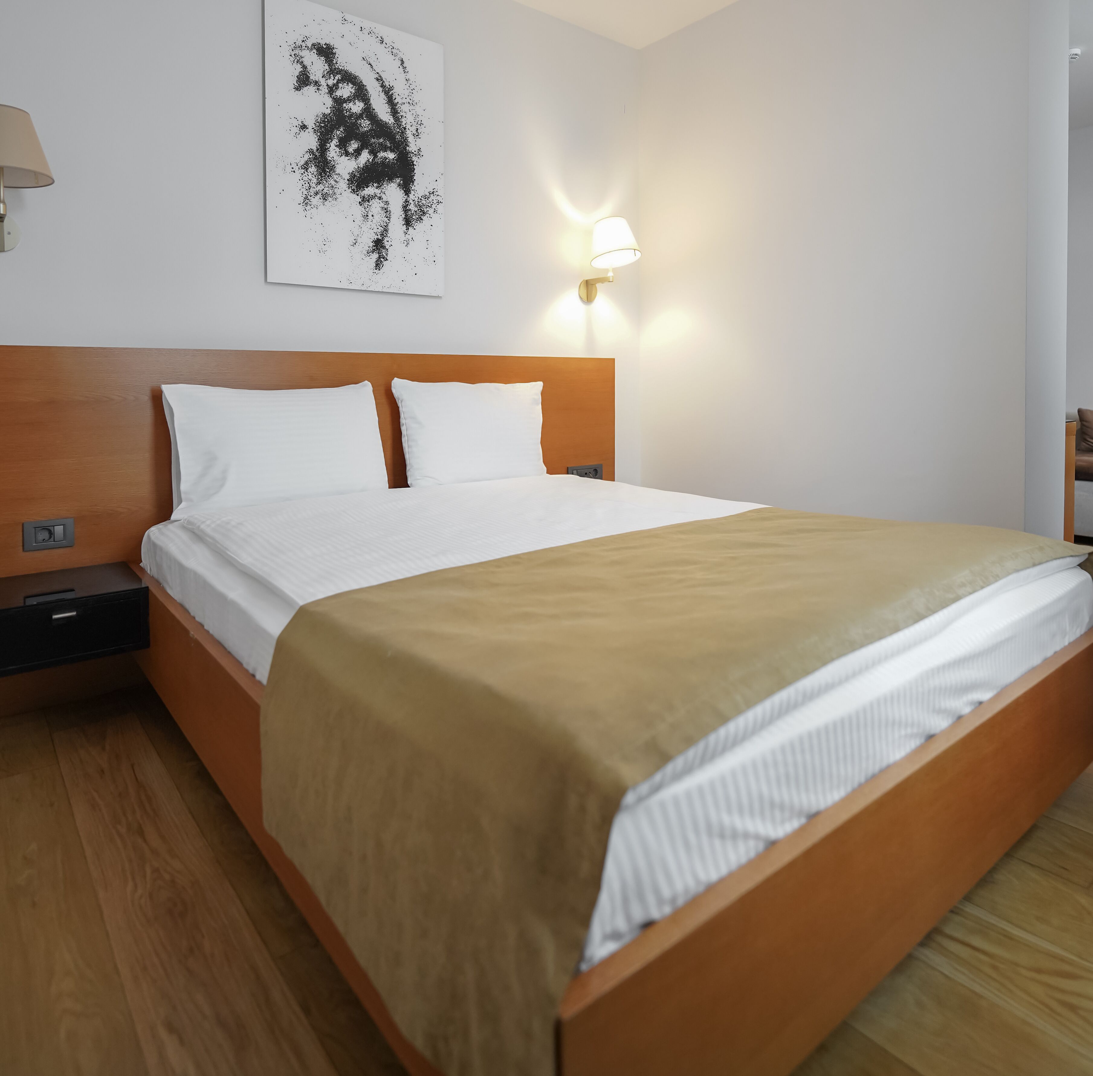 A neatly made bed with white linens and a brown throw, a wooden headboard, a wall-mounted light, and a piece of abstract art above the bed.
