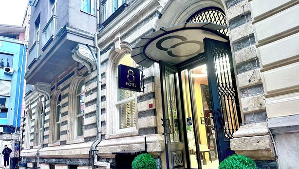 Un edificio histórico con un cartel de hotel visible, grandes ventanales y una entrada imponente con escalones y plantas en macetas.
