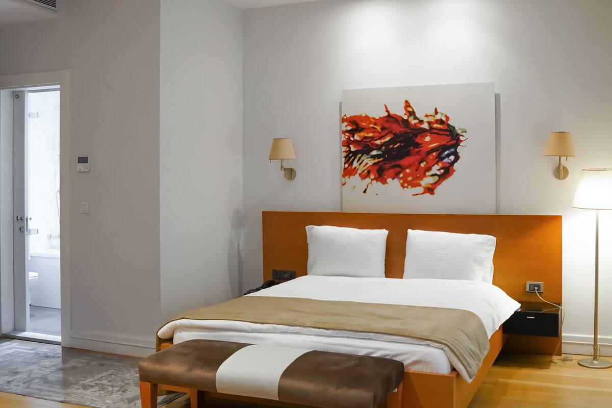 A modern hotel room with a wooden bed, a bench, two bedside lamps, and a colorful abstract painting on the wall.