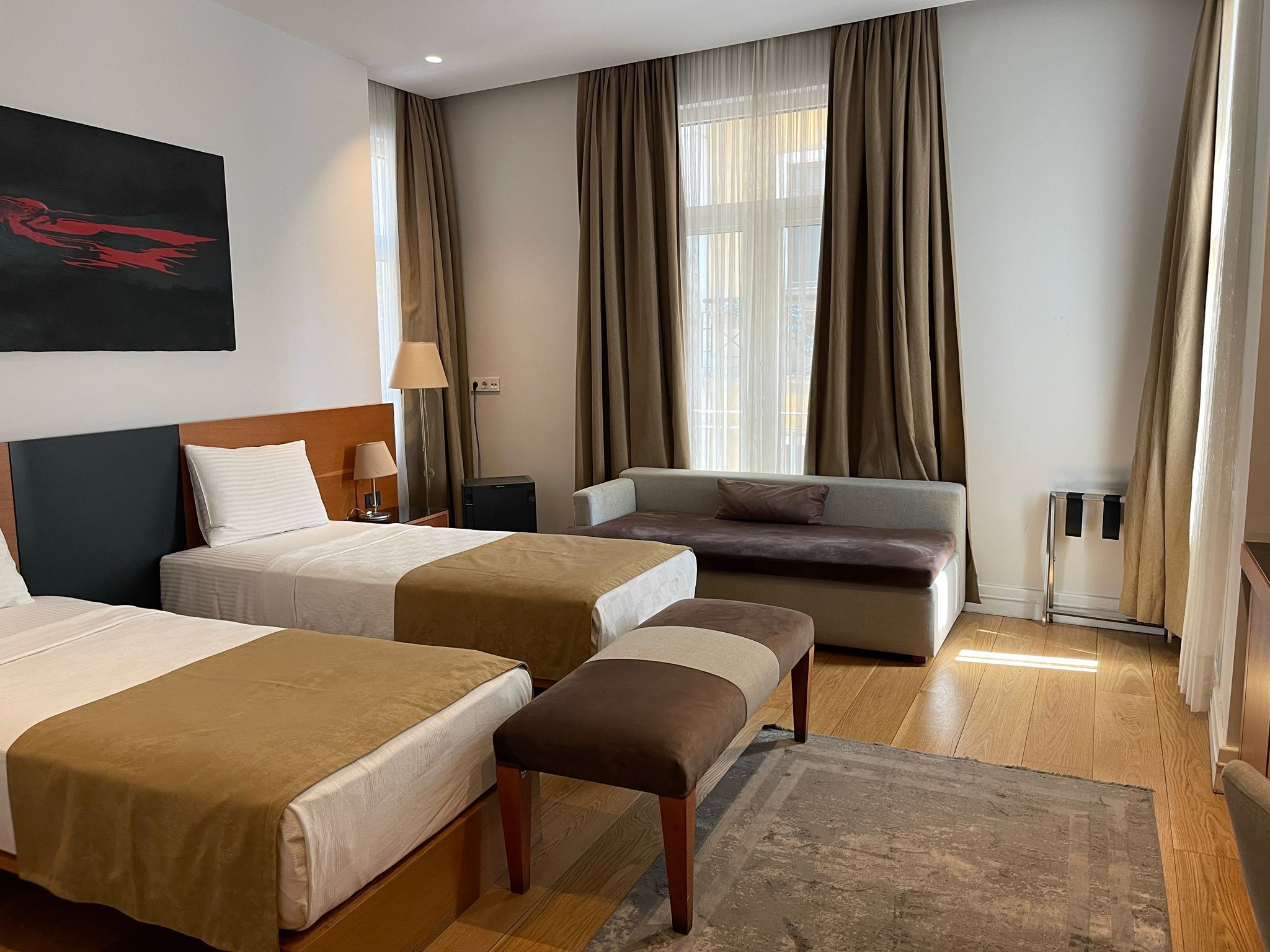 A hotel room with two beds, a sofa, a bench, a lamp, and a large window with curtains.