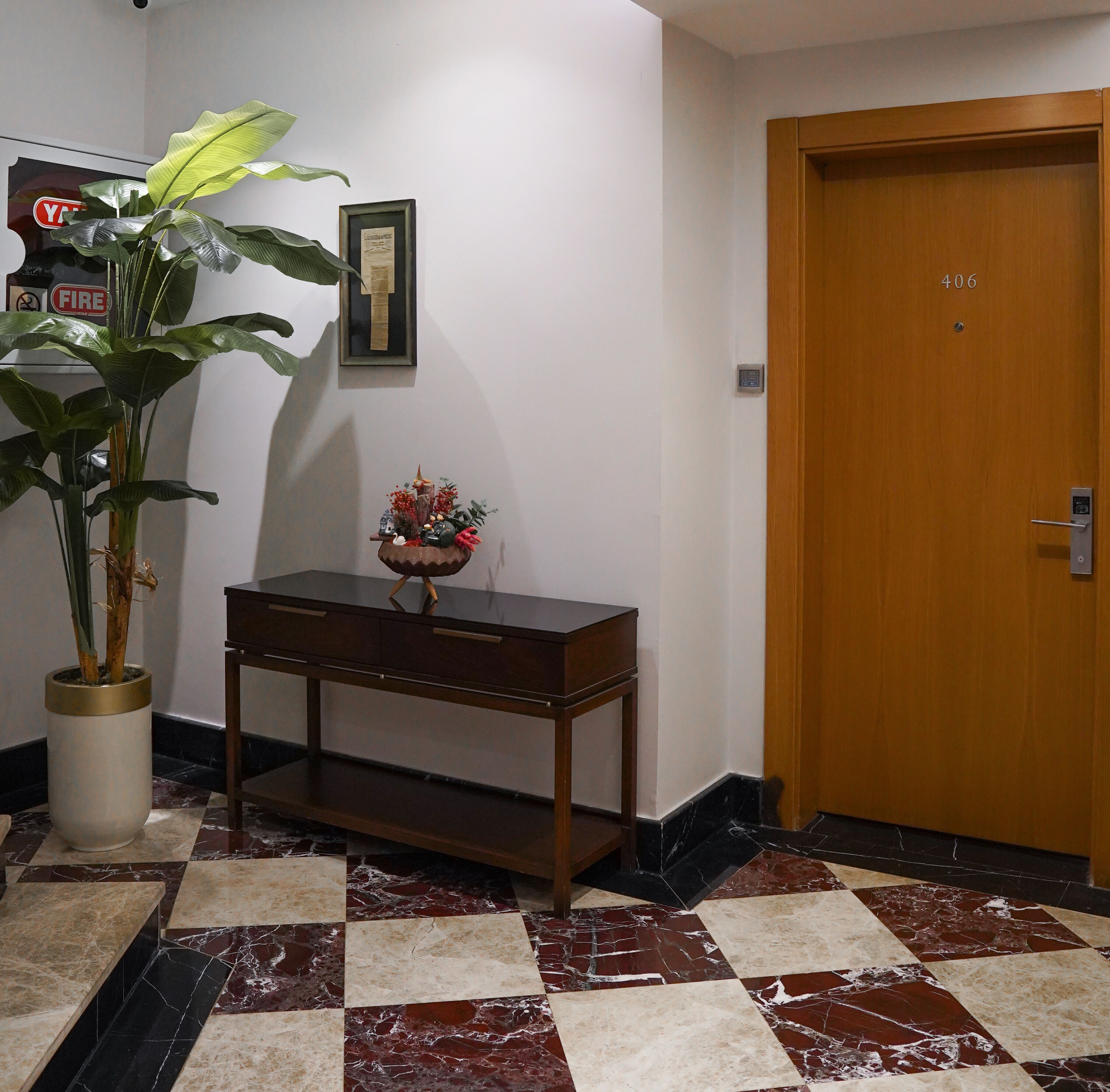 A hallway with a wooden door marked 406, a potted plant, a small table with a vase of flowers, and a framed certificate on the wall.