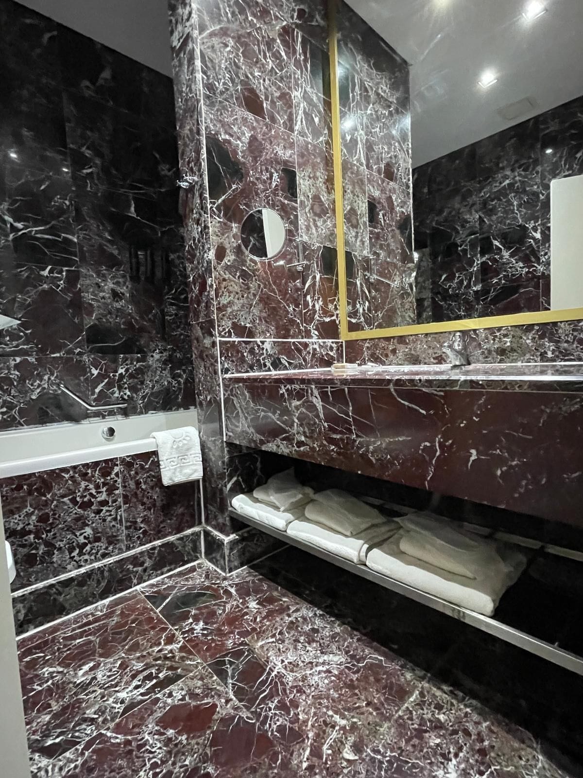 A bathroom with dark marble walls and a gold-framed mirror. There are white towels on a shelf and a white towel hanging on a rack.