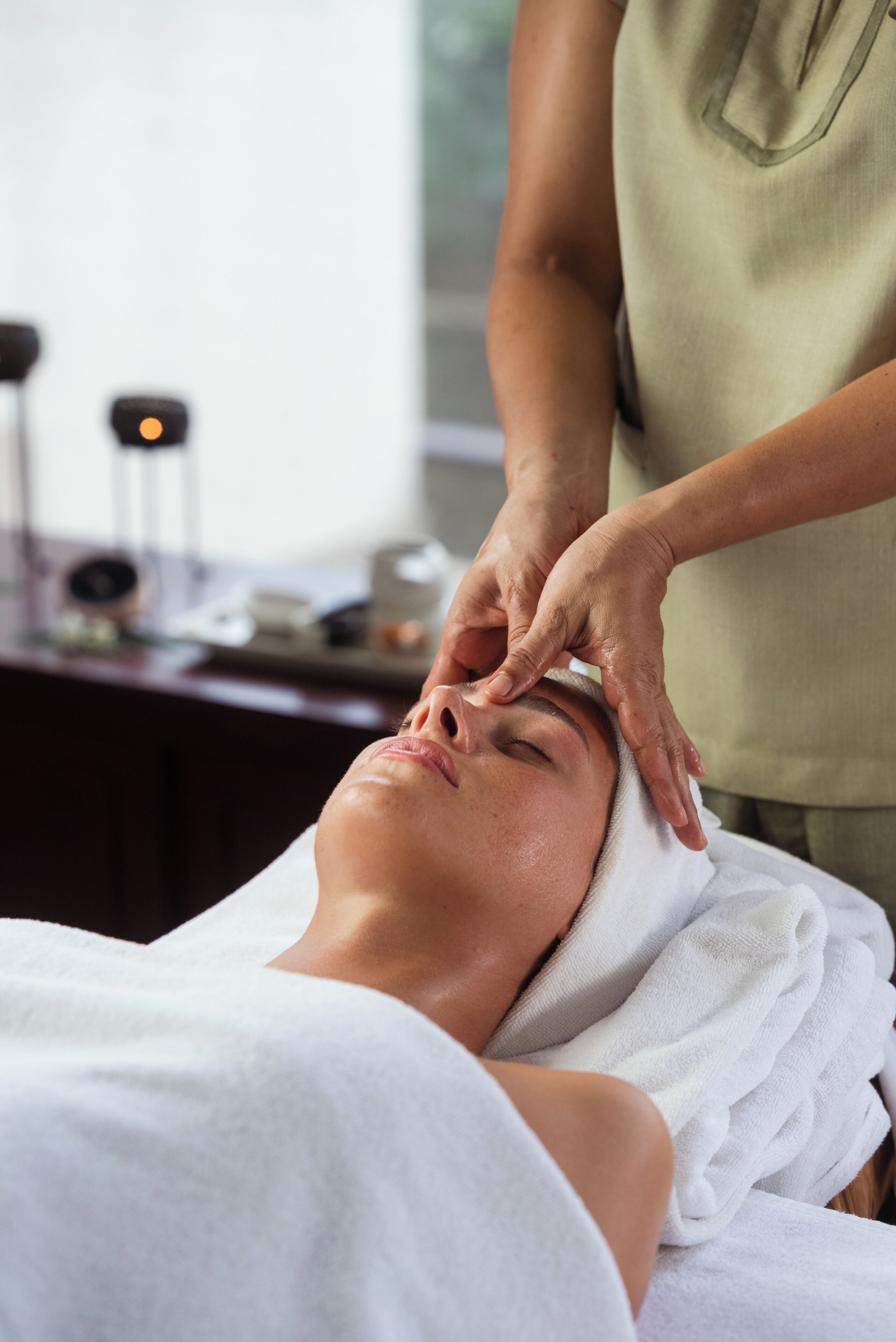 Une personne reçoit un soin du visage dans une pièce qui contient une table de massage, une petite table avec divers objets et une fenêtre en arrière-plan.