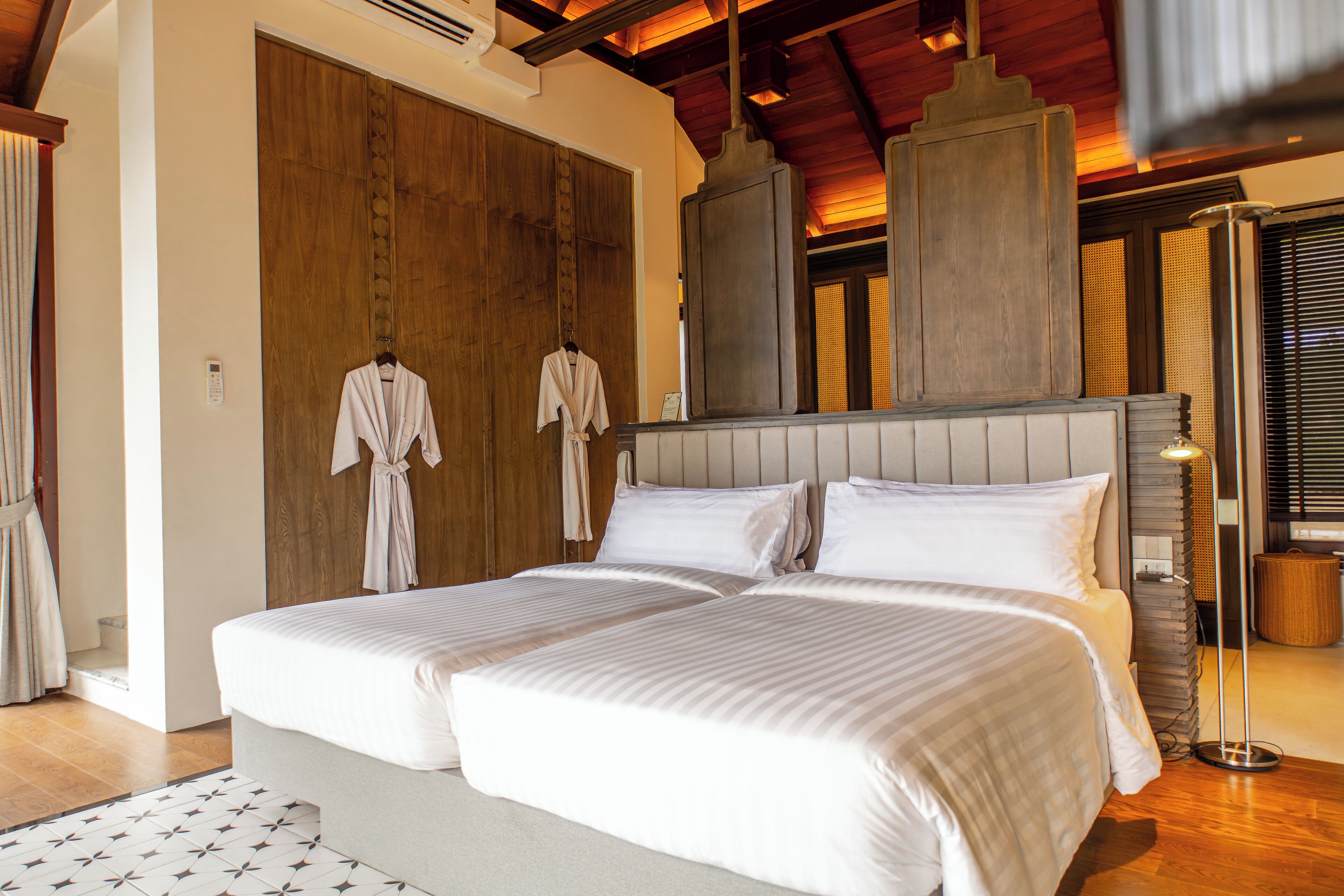 A double bed with a headboard, two robes hanging on a rack, a floor lamp, and a wooden door.