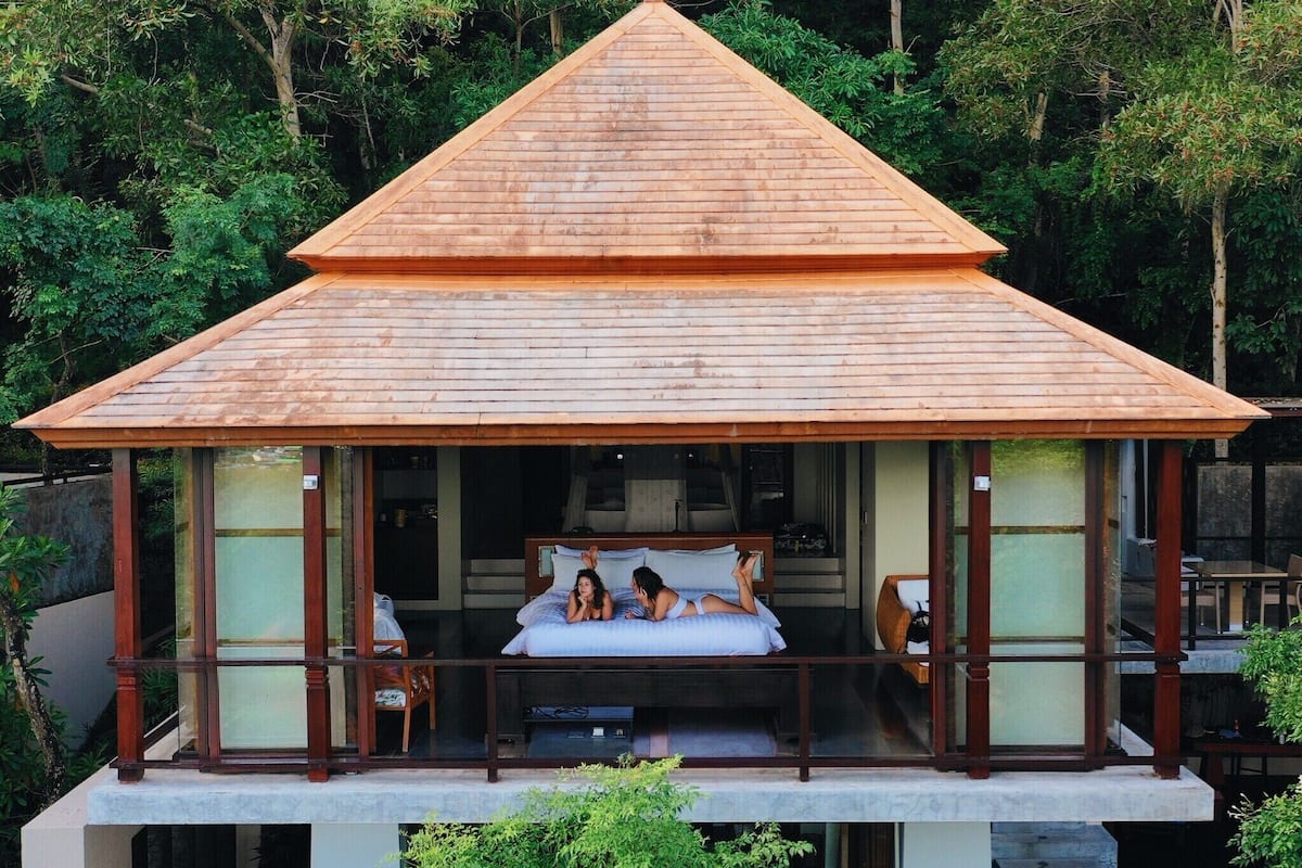 Una estructura de madera de dos pisos con techo inclinado, un balcón con cama y vista a un bosque frondoso.