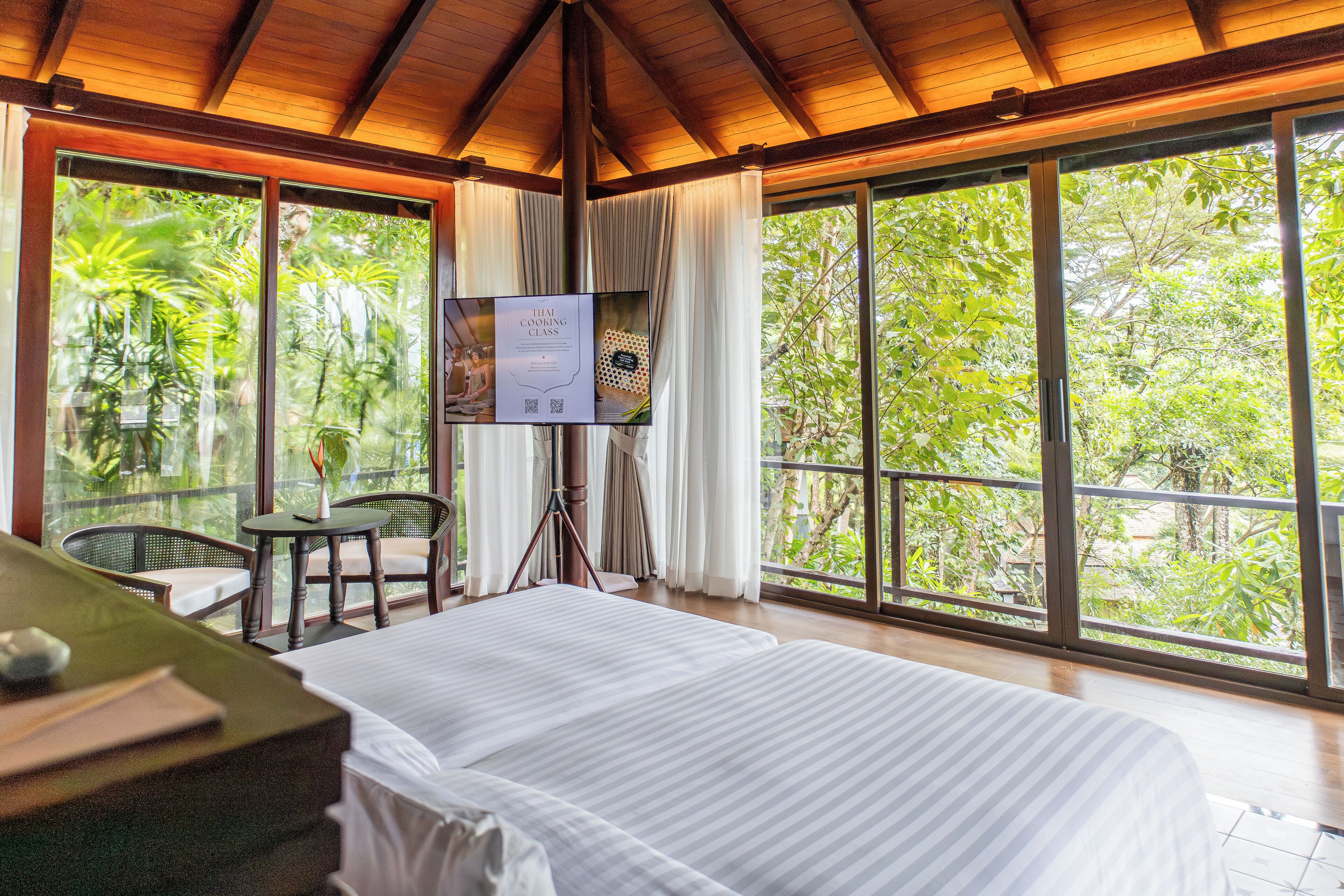 A room with a bed, a table, a chair, and a TV screen displaying a Thai cooking class.