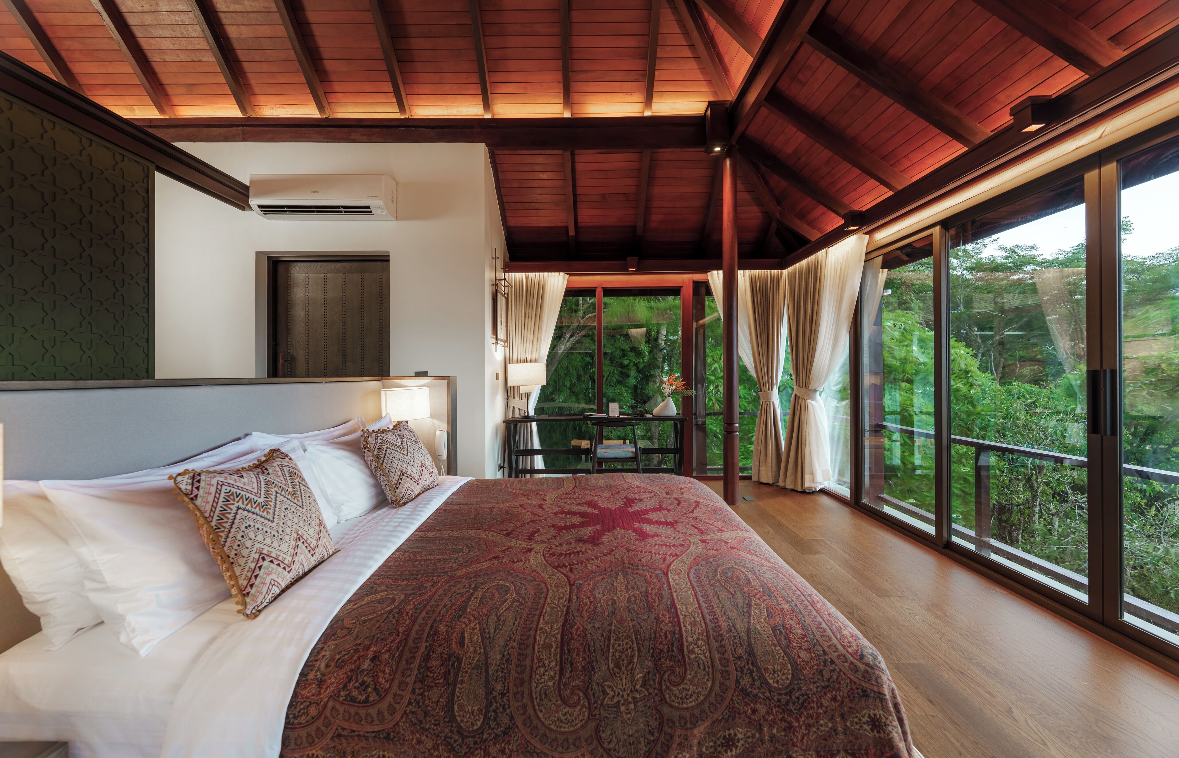 A bedroom with a large bed, a wooden ceiling, and a view of greenery outside.