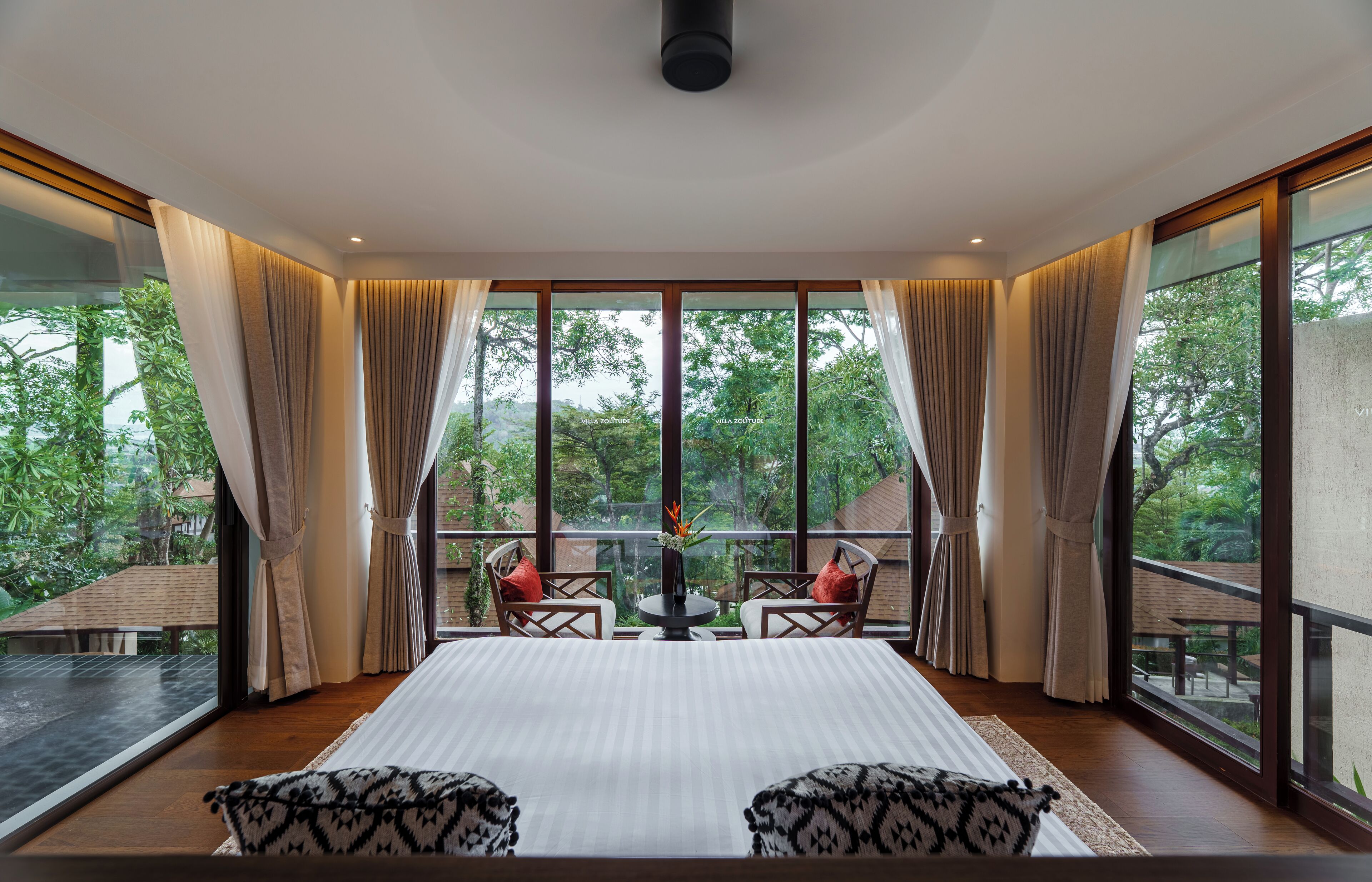 A spacious room with a large bed, two chairs, and a table, featuring large windows and curtains.