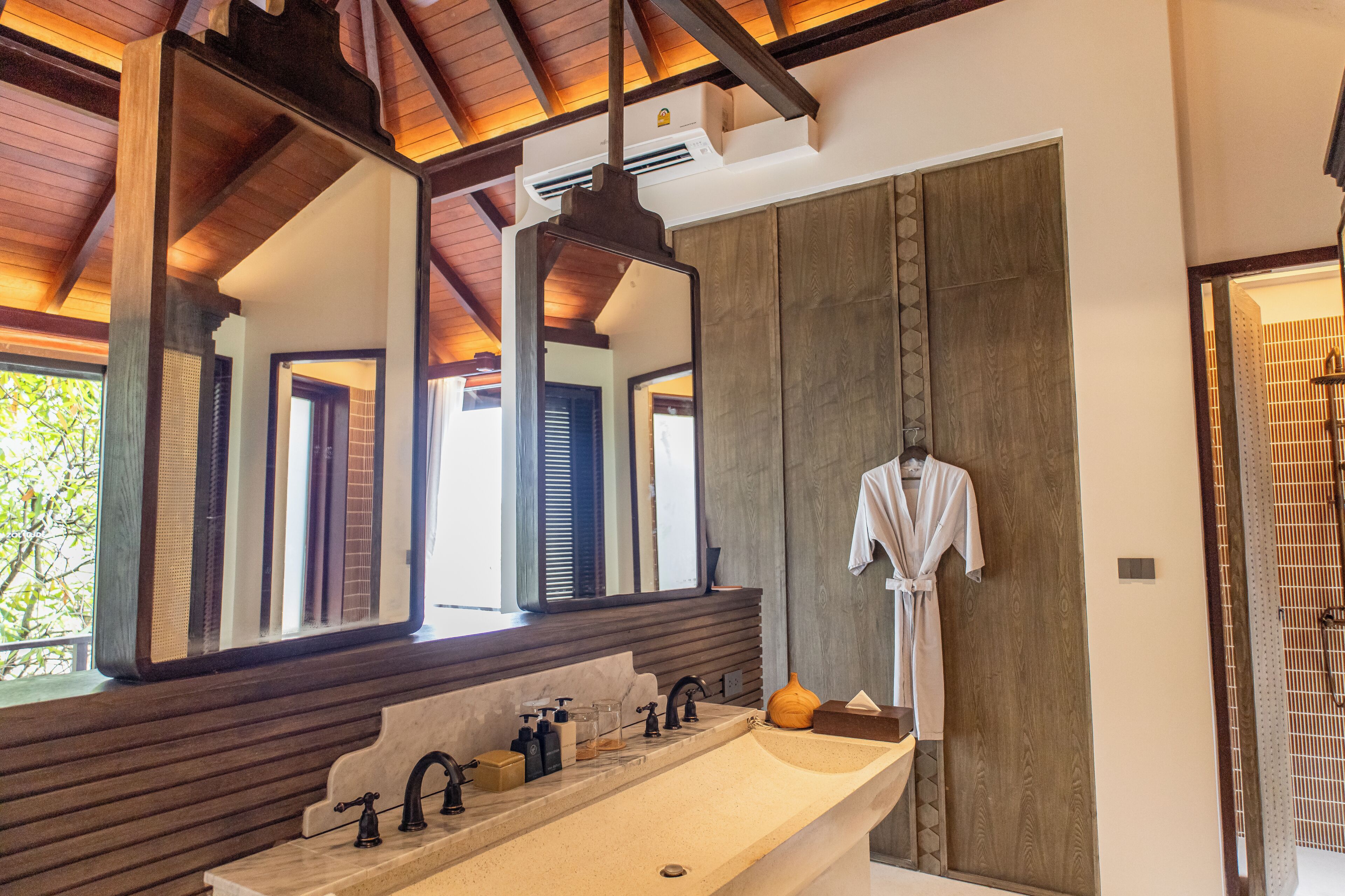 A bathroom with a large mirror, wooden ceiling beams, a long sink, and a robe hanging on the wall.