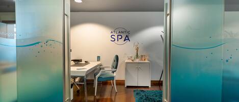 A spa room with a glass partition, a desk, a chair, a cabinet, and a blue rug.