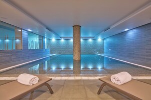 Piscina interior con pared de azulejos, dos sillas de descanso con toallas y un pilar central.