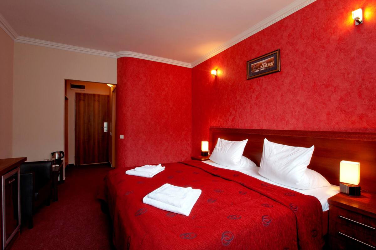 A hotel room with a red carpet, a wooden headboard, a bed with white linens, two bedside lamps, and a framed picture on the wall.