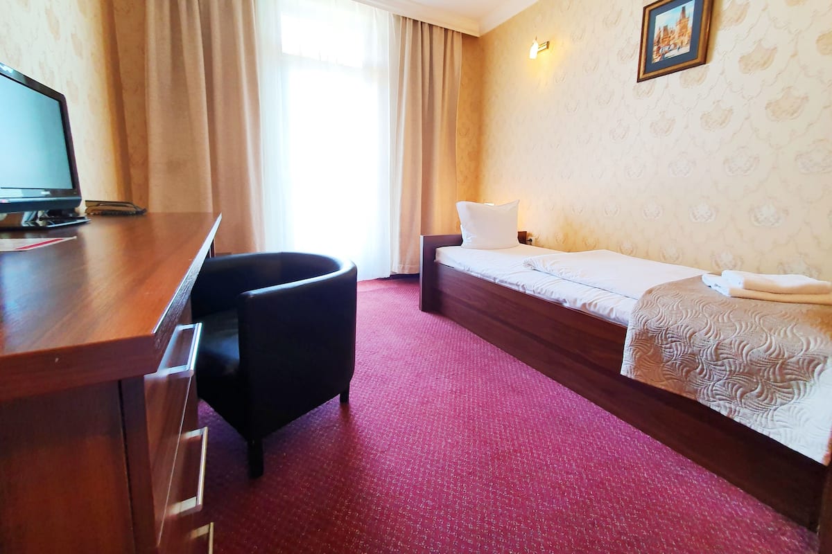 A hotel room with a wooden desk, a black armchair, a single bed with a white comforter, and a patterned wall.