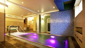 Uma piscina interior moderna com um elemento aquático, cercada por paredes com azulejos em mosaico e uma escadaria que leva a um nível superior.