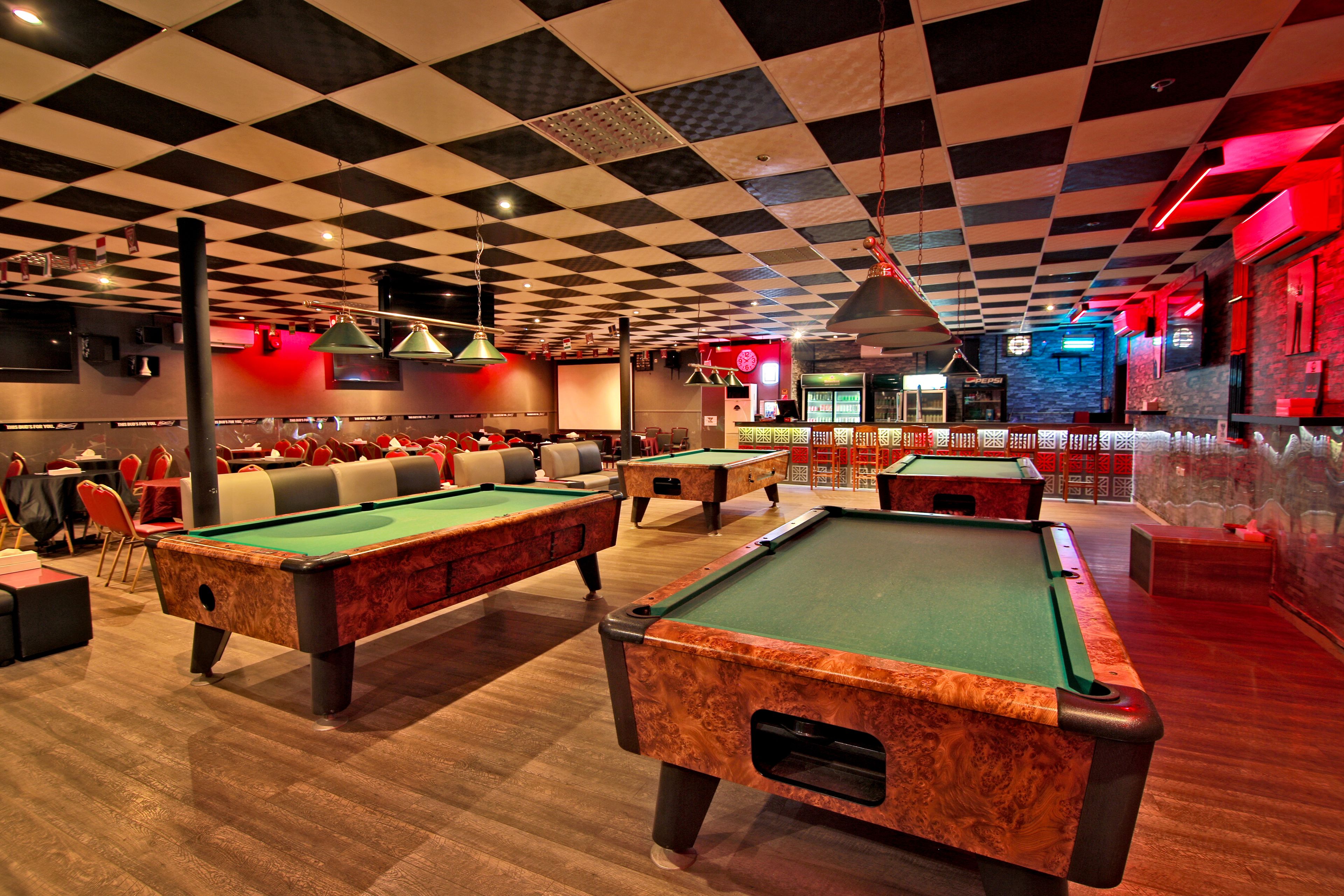 Una sala de billar con varias mesas de billar, techo a cuadros rojos y negros, y una zona de bar con luces de neón.