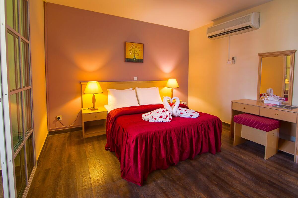 A hotel room with a red bedspread, two bedside lamps, a wooden desk with a mirror, and a wall-mounted air conditioner.