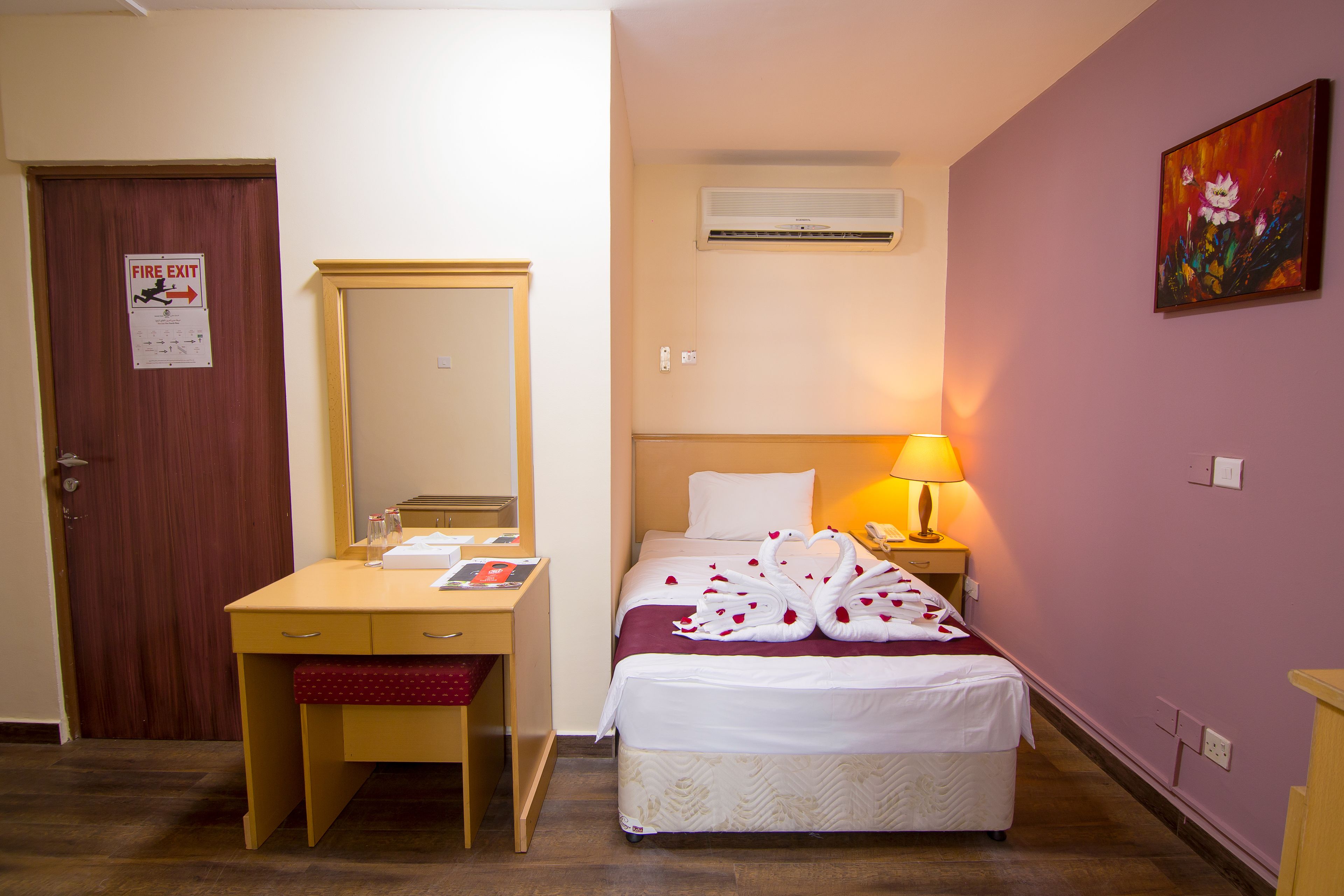 Quarto de hotel com cama, escrivaninha, espelho, abajur e sinal de saída de incêndio.