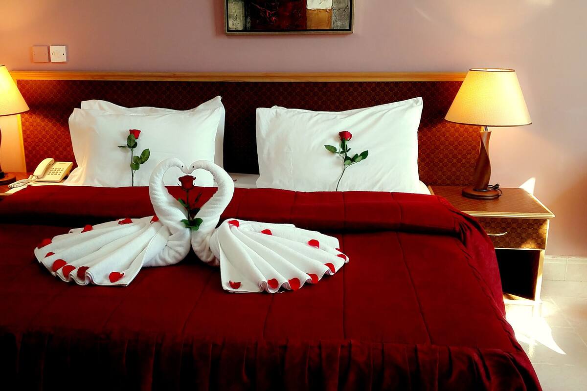 A hotel room with a bed, red bedding, two bedside lamps, a telephone, and a swan-shaped towel arrangement with roses.