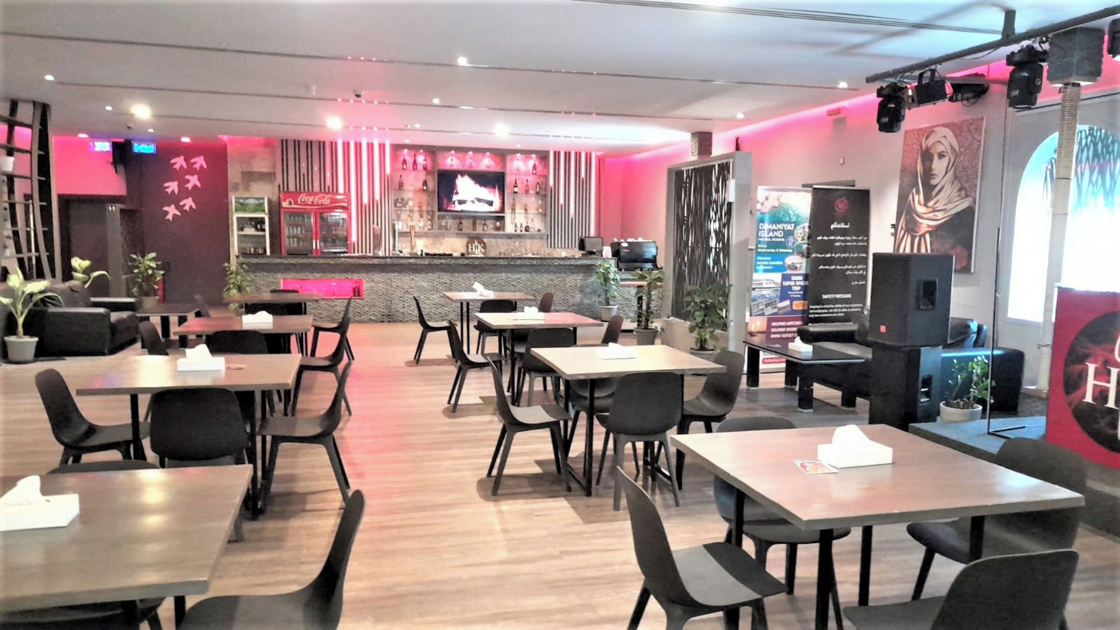 Un restaurant moderne avec des tables en bois et des chaises noires, un coin bar avec un réfrigérateur rempli de Coca-Cola et une grande affiche accrochée au mur.