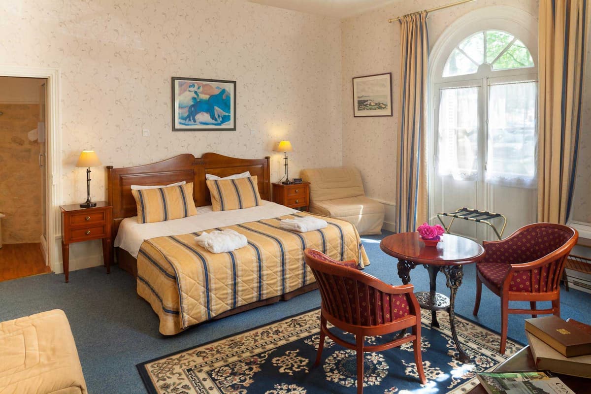 Ein Hotelzimmer mit einem großen Bett, zwei Nachttischen mit Lampen, einem Holzstuhl, einem kleinen runden Tisch mit einer Vase und Blick ins Freie durch ein Fenster.
