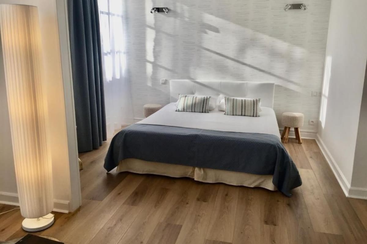 A modern bedroom with a large bed, a bedside table, a floor lamp, and a view of the sky through a window with curtains.