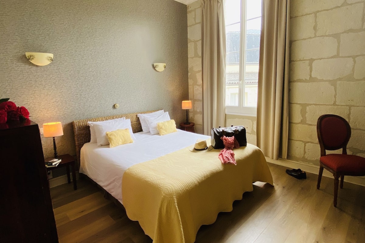 Ein Hotelzimmer mit Bett, Nachttischen, einem Sessel, einem Fenster mit Vorhängen und einer Steinwand.