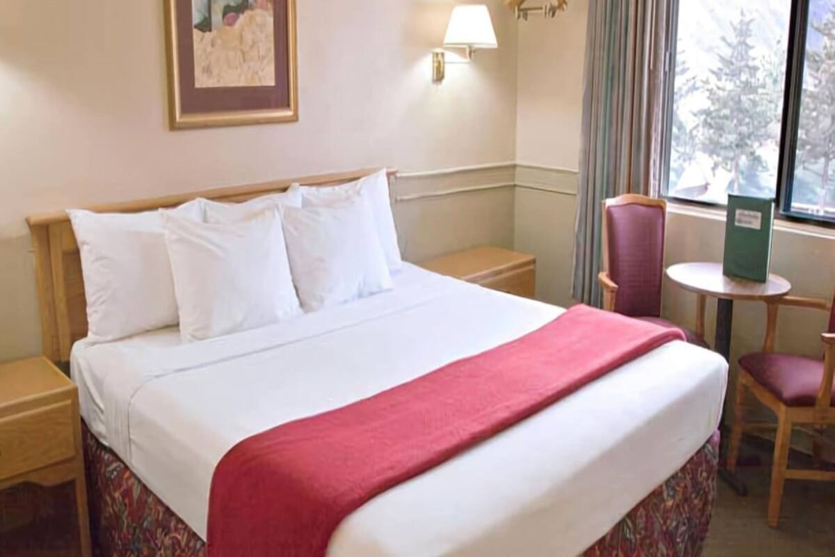 Une chambre d’hôtel avec un grand lit, une table de chevet, une chaise, une petite table, une lampe et une fenêtre donnant sur des arbres.