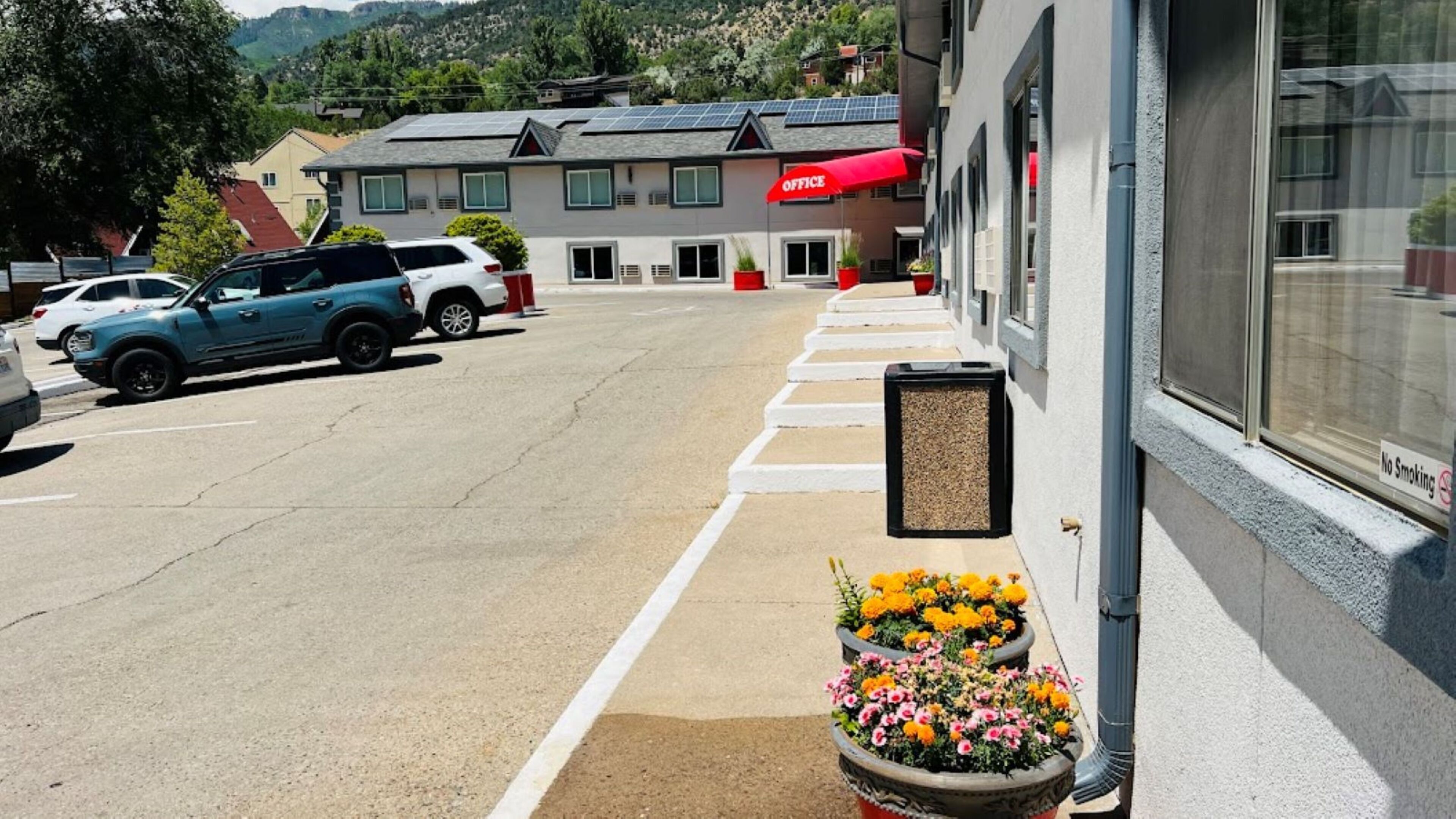 A parking lot with a building labeled "OFFICE" in the background, a no smoking sign, and flower planters on the sidewalk.