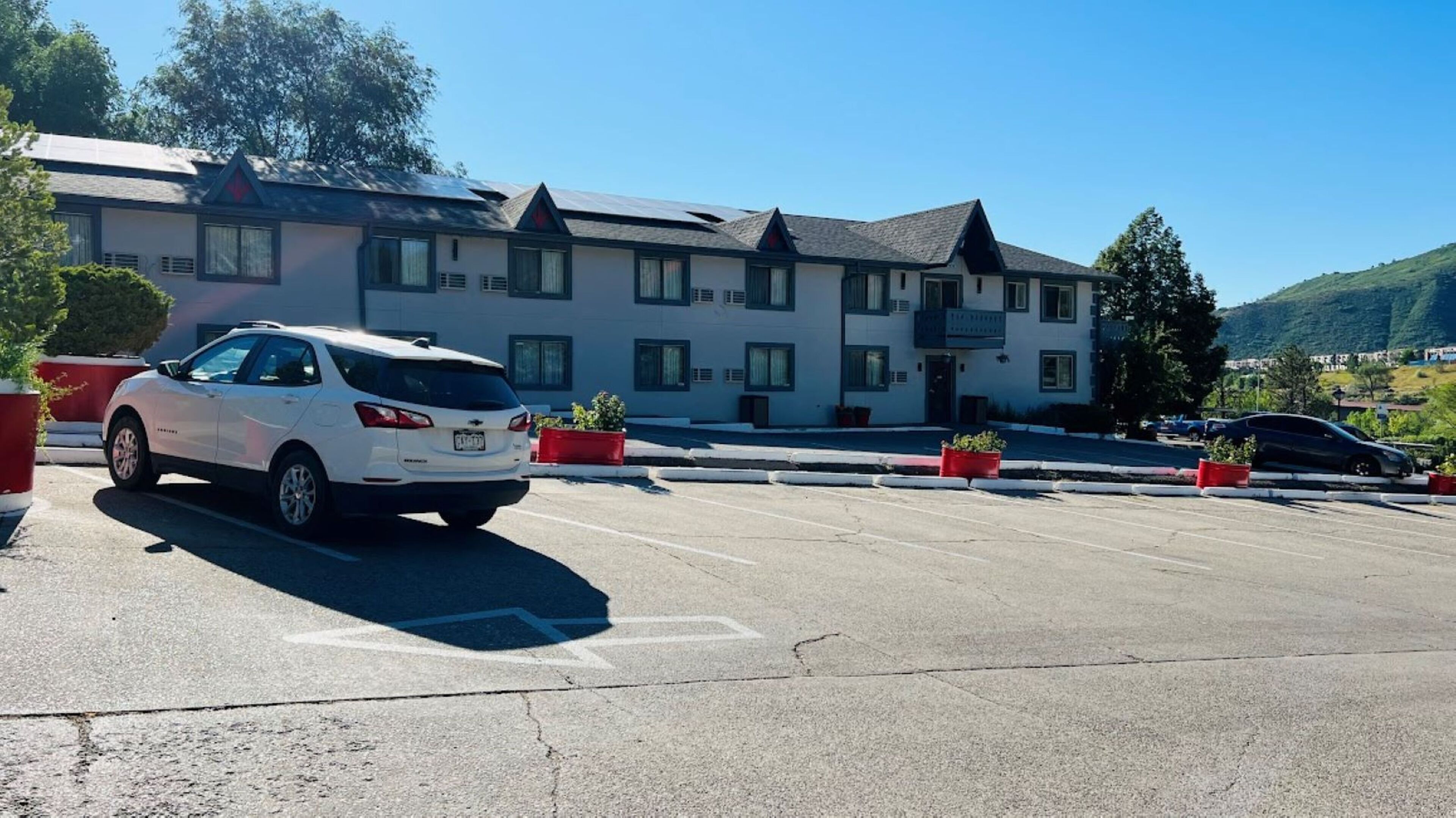 A parking lot with a white car parked in front of a building with multiple windows and a sloped roof.