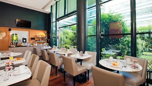 A modern dining area with round tables and chairs, a kitchen in the background, and large windows offering a view of greenery. - Residhome Monceau (Bois-Colombes)