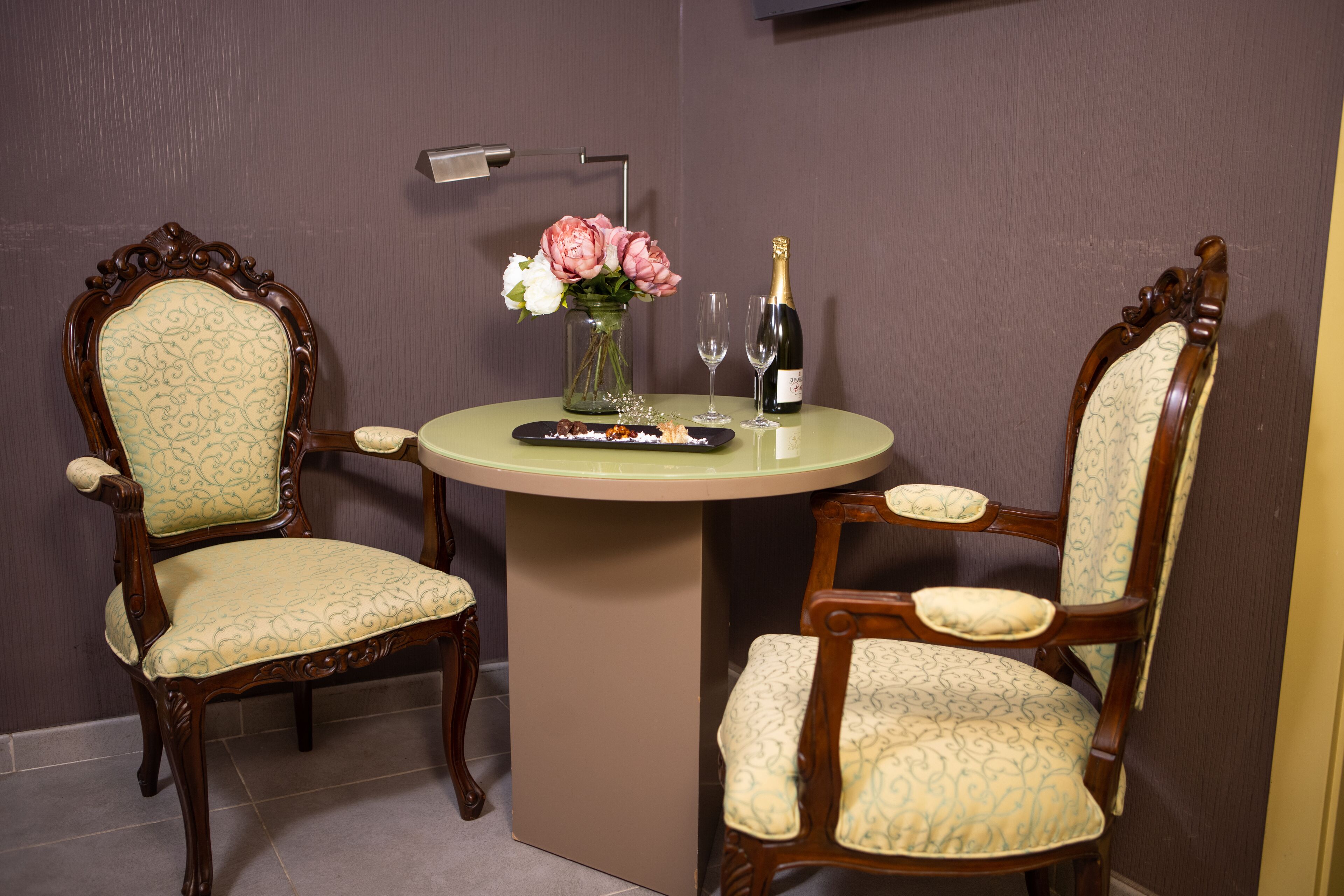A room with two ornate wooden chairs with patterned upholstery, a round table with a vase of flowers, a bottle of champagne, and two glasses.