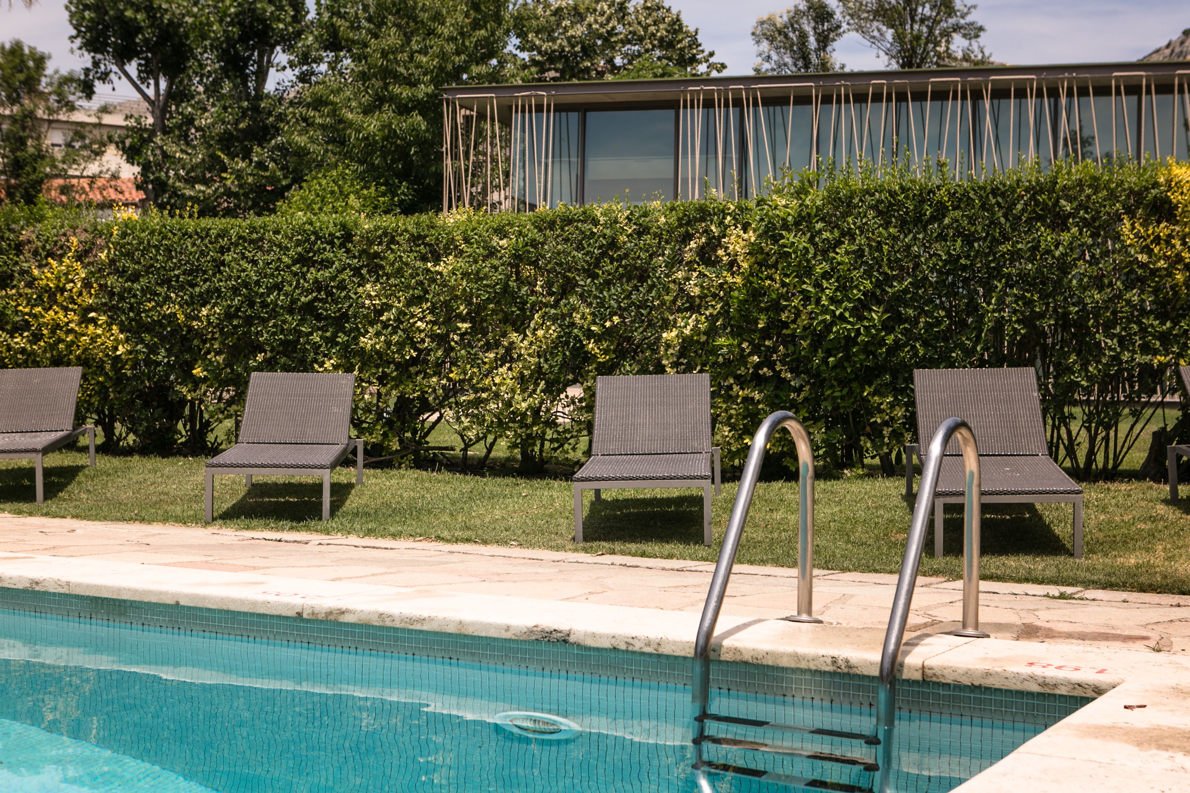 Une piscine avec un escalier en métal, entourée de chaises longues et d’une haie.