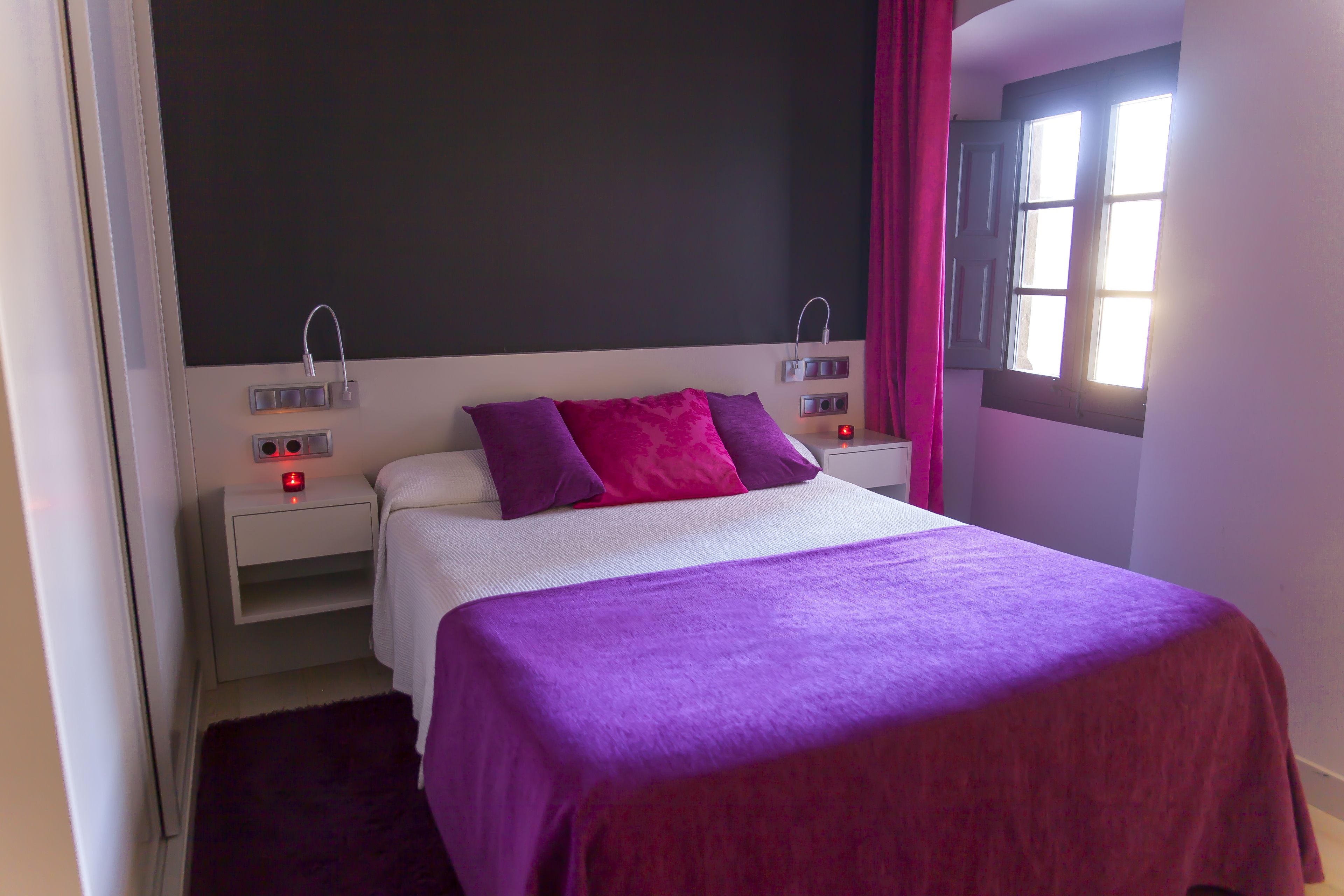 A bedroom with a bed, two bedside tables, a nightstand, a window with curtains, and a wall-mounted light fixture.