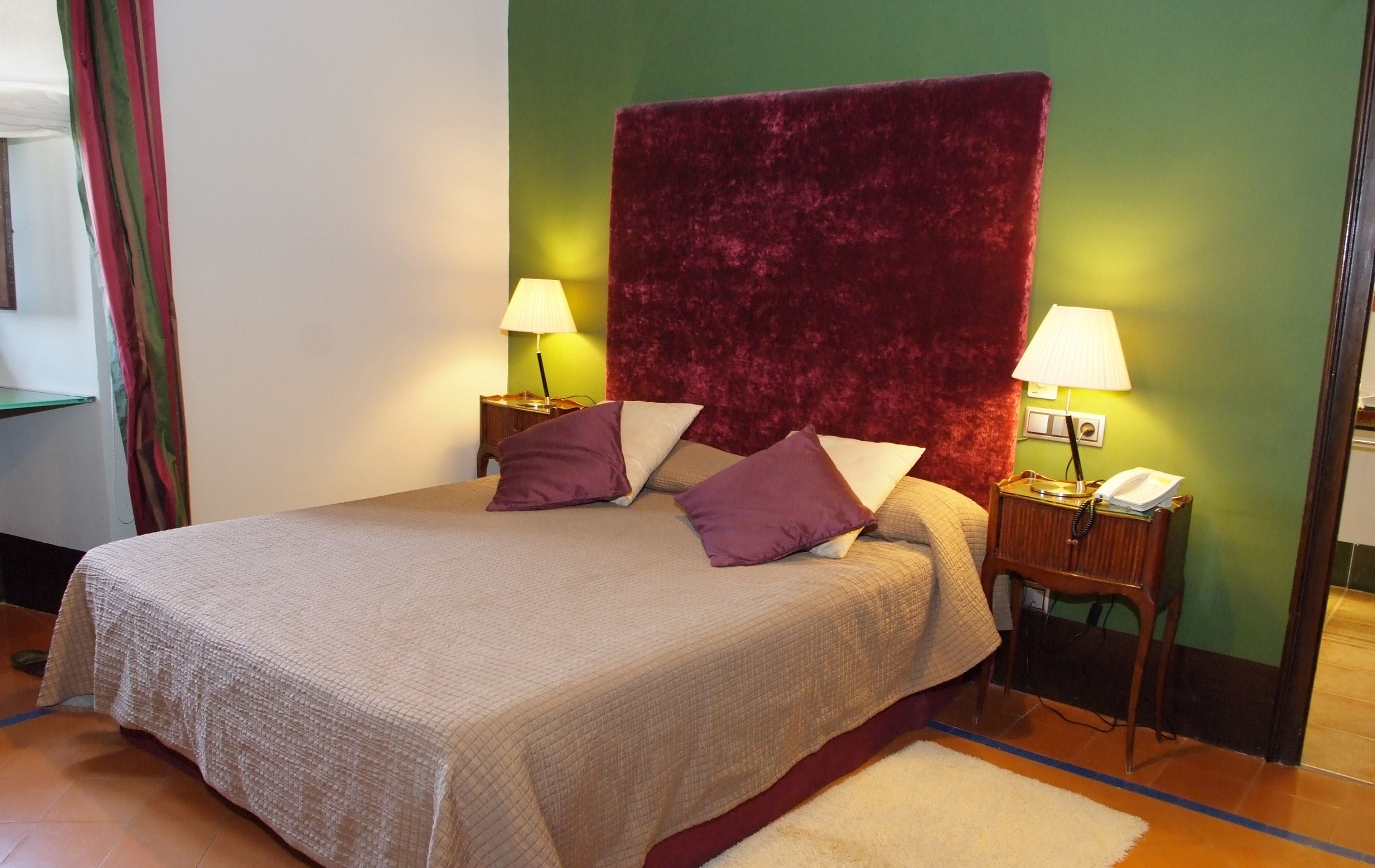 Una cama bien tendida con un amplio cabecero rojo, dos lámparas de noche y un teléfono sobre una mesita de madera.