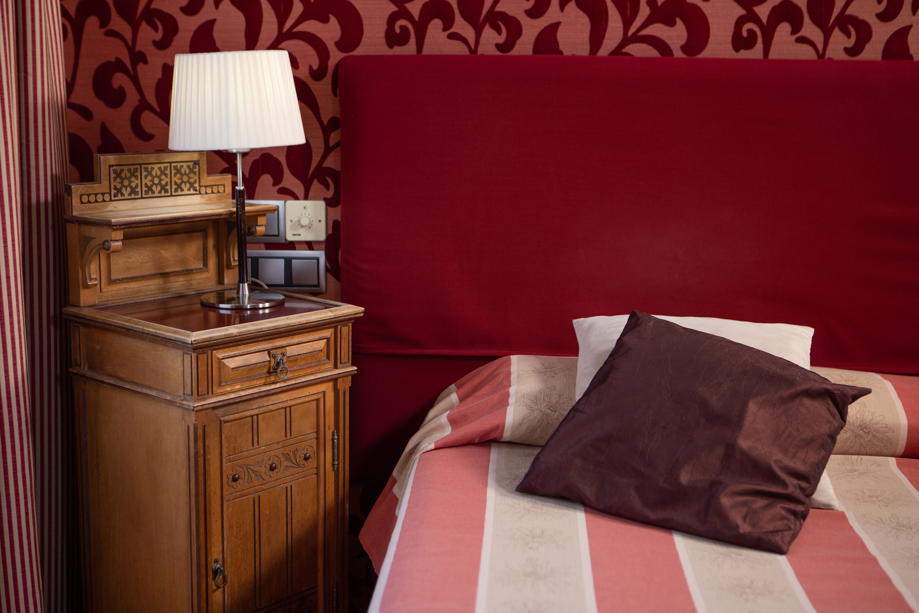 Una cama bien hecha con una colcha a rayas y dos almohadas, una mesita de noche de madera con una lámpara, y una pared con papel tapiz rojo estampado.