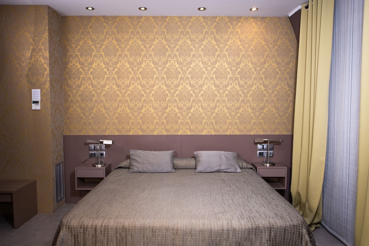 A hotel room with a patterned wallpaper, a large bed with two pillows, two bedside tables with lamps, and a window with curtains.