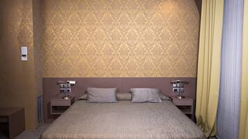 A hotel room with a patterned wallpaper, a large bed with two pillows, two bedside tables with lamps, and a window with curtains.