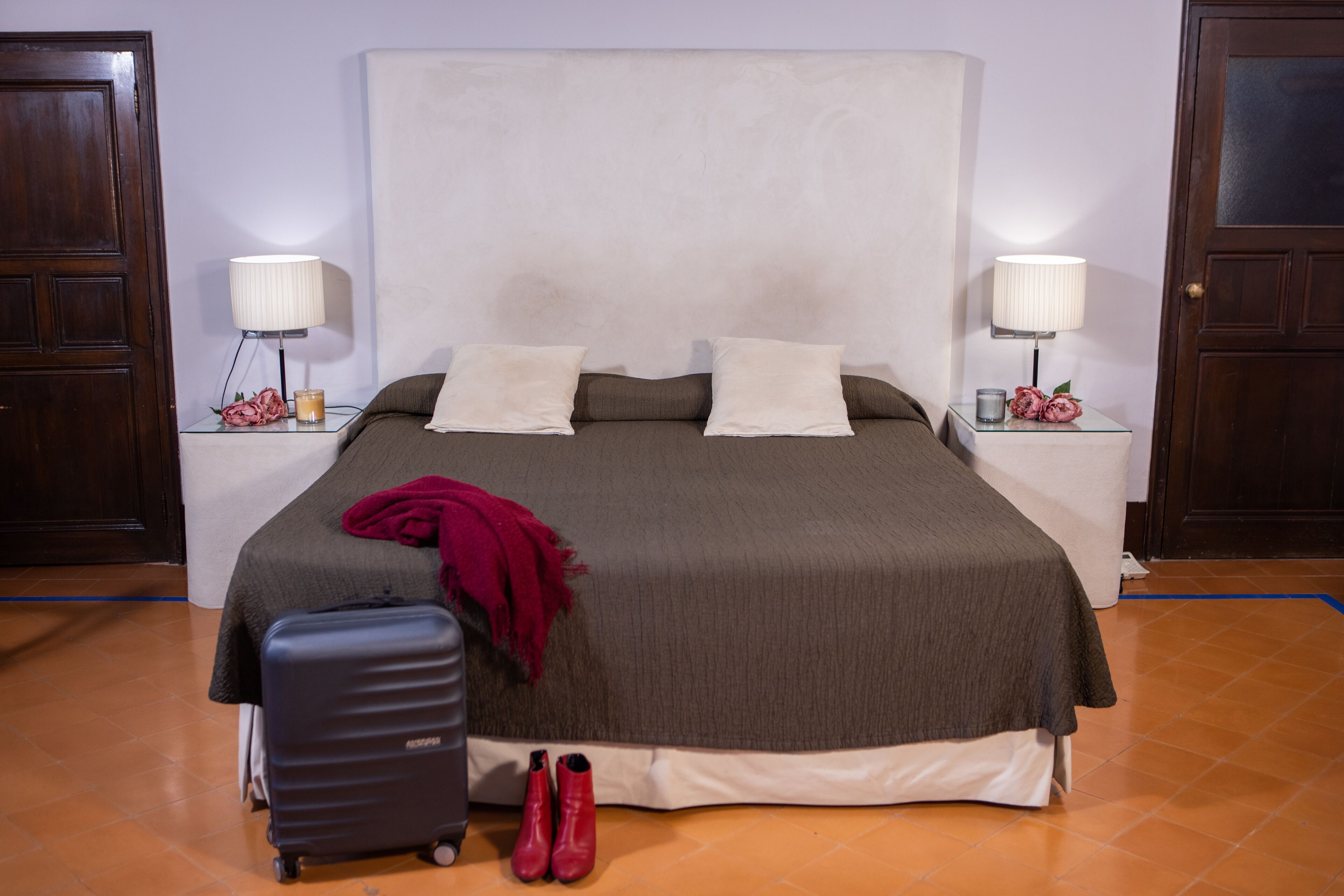 Un letto rifatto con un piumone grigio e bianco, due cuscini, una coperta rossa, una valigia e un paio di scarpe rosse sul pavimento.