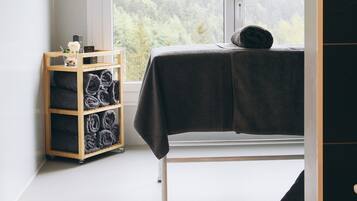A bedroom with a large window, a bed with a dark blanket, a wooden shelf with towels, and a view of a forested area.