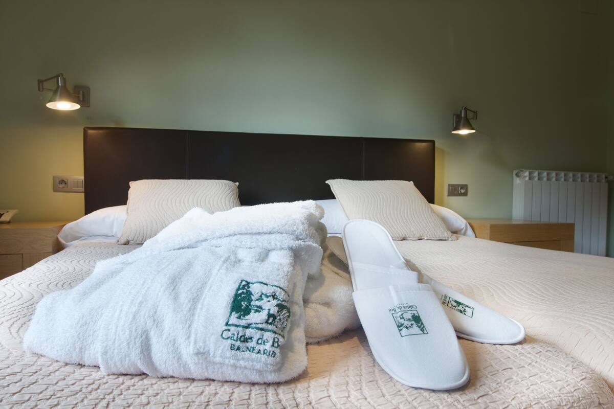 Cama bem arrumada com lençóis brancos e chinelos, cabeceira e duas luminárias fixadas na parede.
