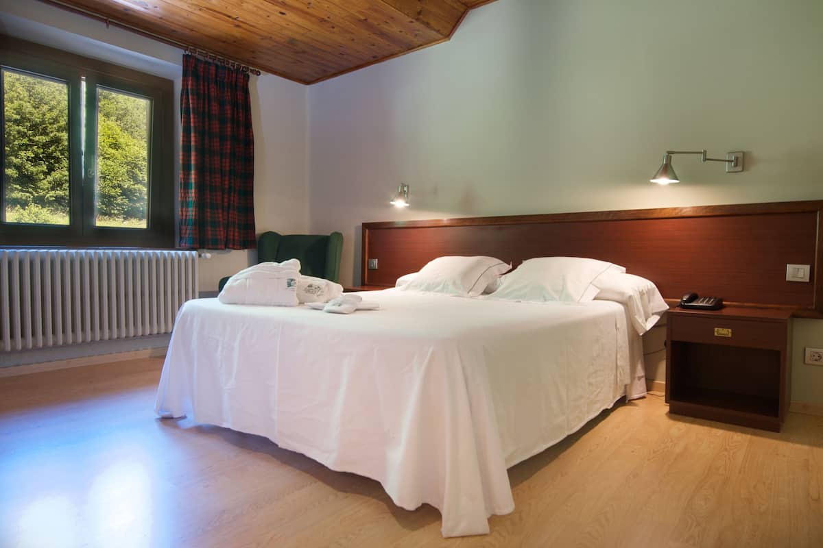 Cama de casal com lençóis brancos, mesa de cabeceira com abajur e vista para árvores pela janela.