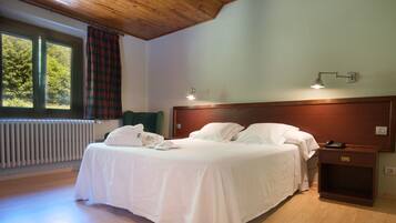 Cama doble con sábanas blancas, mesita de noche con lámpara y vista a árboles por la ventana.