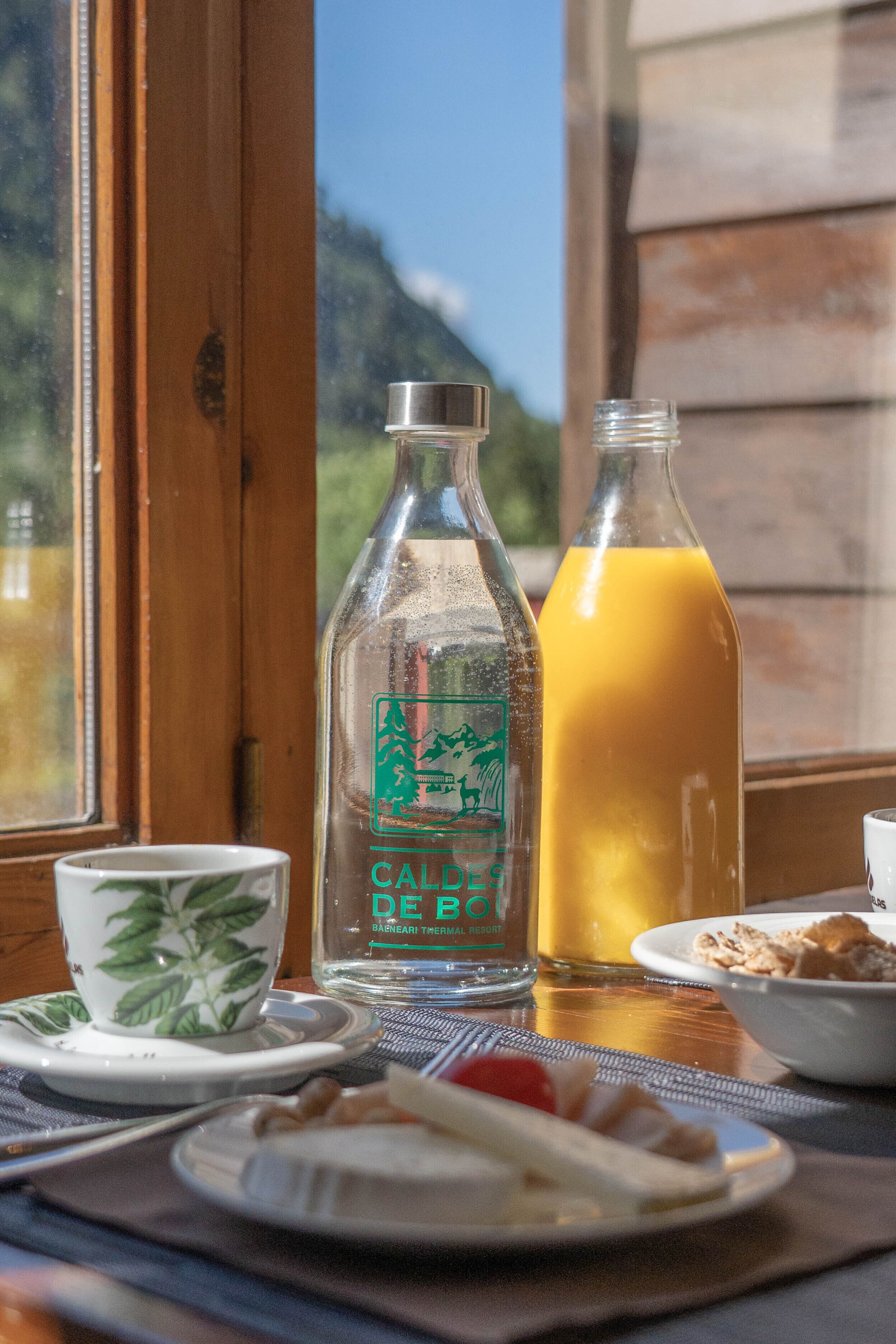 A breakfast table with a bottle of Caldes de Boi, a glass of orange juice, and a bowl of cereal.