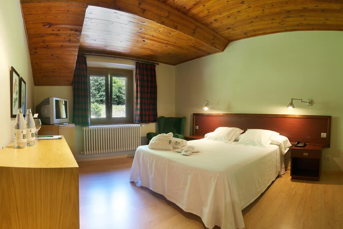 Quarto de hotel com teto de madeira, cama com lençóis brancos, mesa de cabeceira com abajur, janela com cortinas xadrez e radiador.