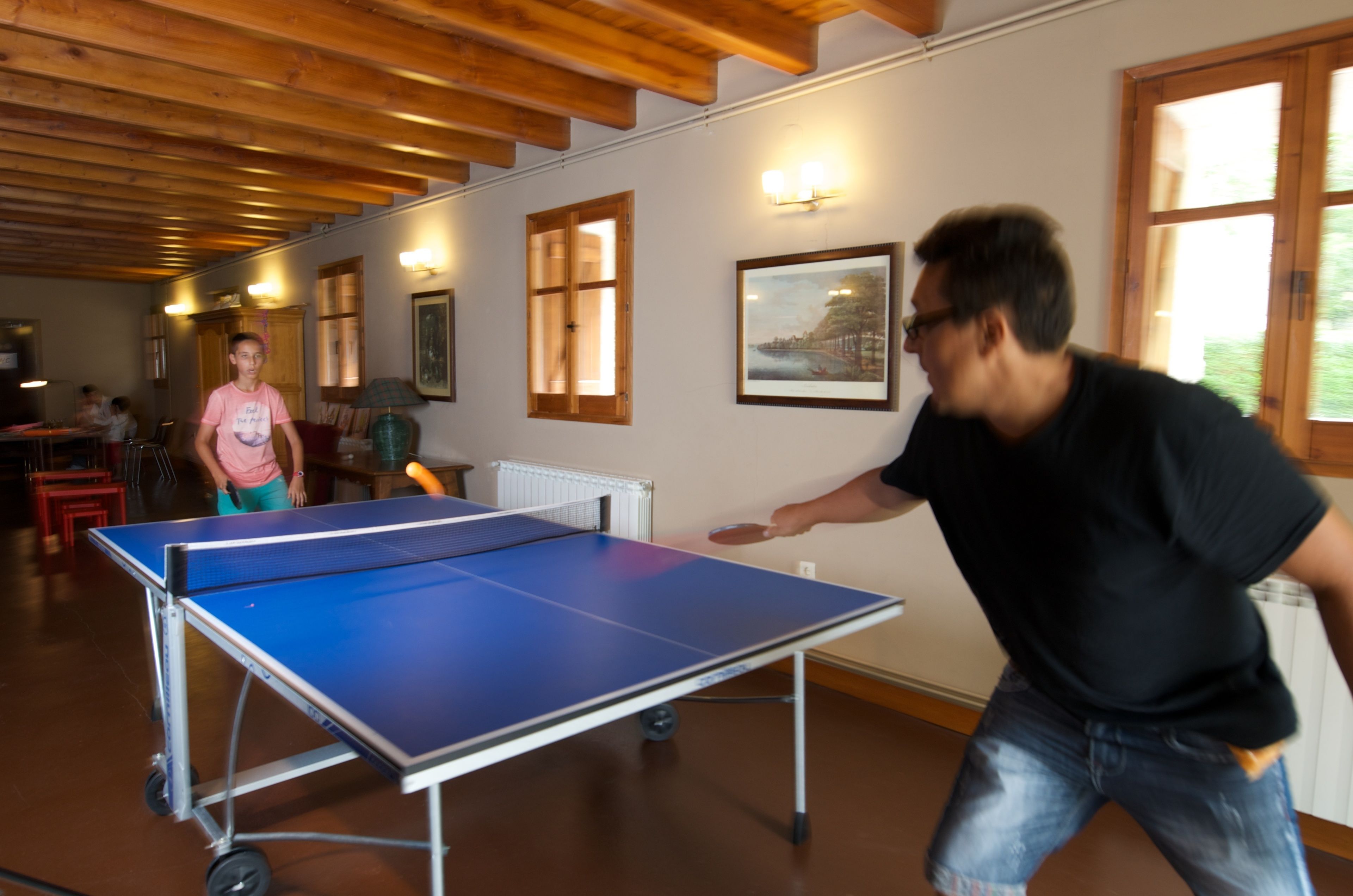 A ping pong table in a room with wooden beams on the ceiling, a radiator, and framed pictures on the wall.