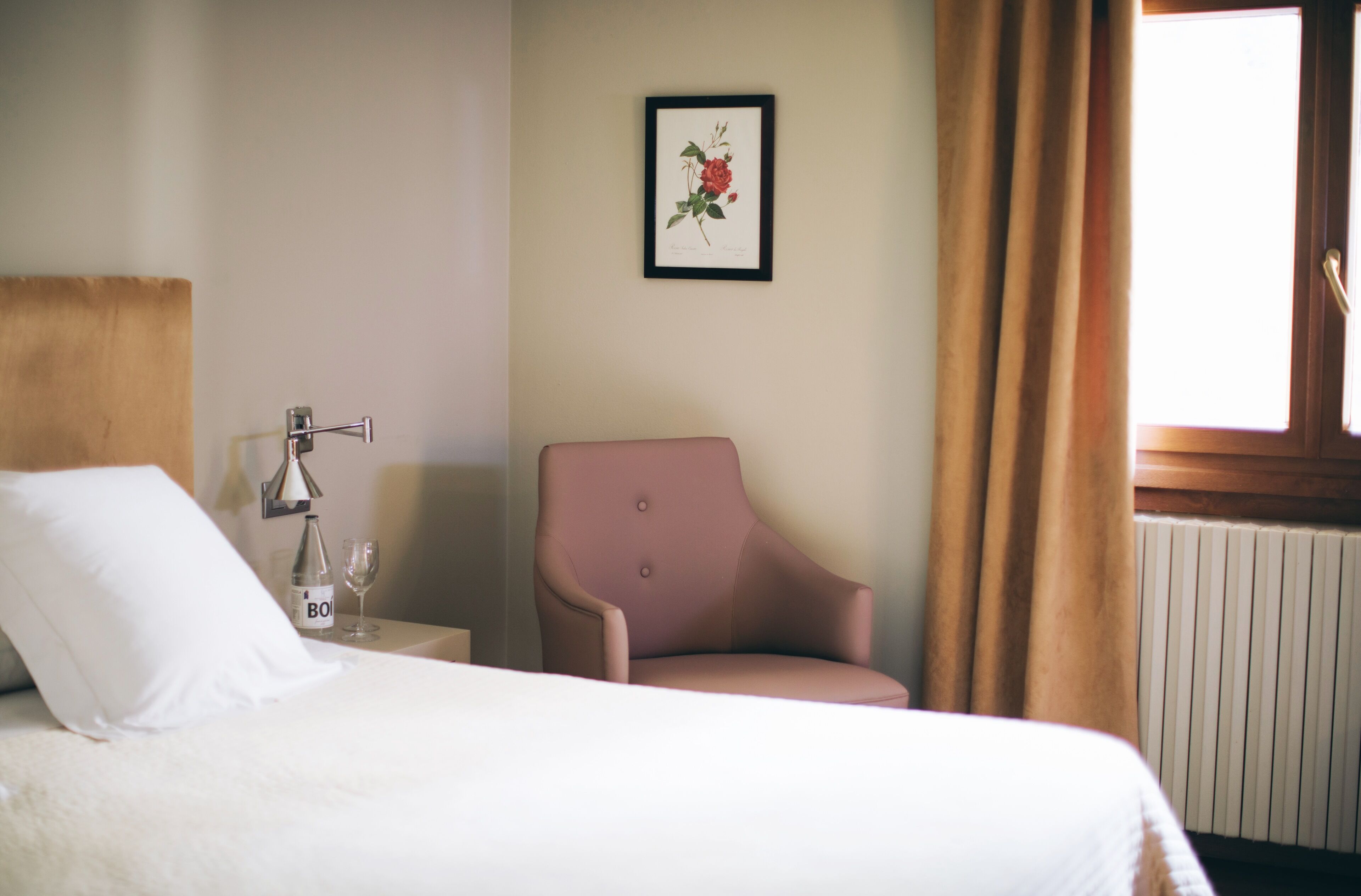 Habitación de hotel con cama, una silla, una mesita de noche con una botella y un vaso, e impresión enmarcadas de motivos botánicos en la pared.