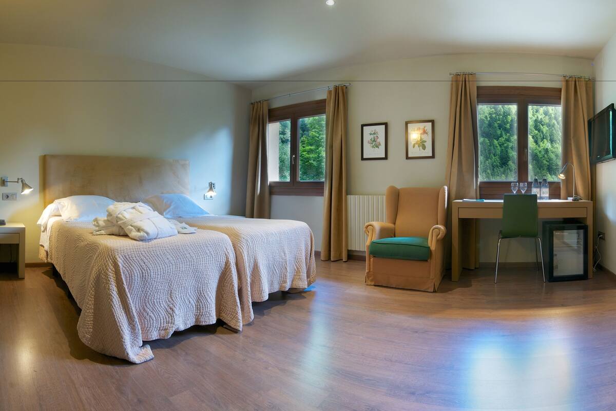 Quarto de hotel com duas camas, uma mesa, uma cadeira e vista para o exterior através de amplas janelas.