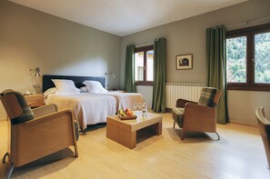 Habitación de hotel con una cama, dos sillas, una mesita con fruta y copas de vino, y una ventana con cortinas.